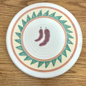 May include: White ceramic trivet with a green, yellow, and orange border. The center of the trivet features two red chili peppers.