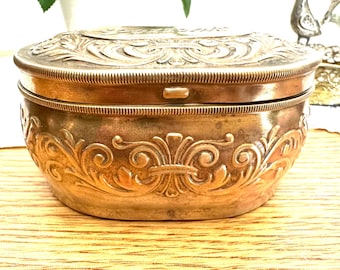 May include: An antique, oval-shaped, gold-toned metal box with ornate floral and scrollwork detailing. The box has a hinged lid and a textured rim. The box is sitting on a wooden surface.