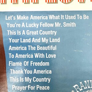 Op de afbeelding: Een vinylplatenhoes met een blauwe achtergrond en een lijst met songtitels: Let's Make America What It Used To Be, You're A Lucky Fellow Mr. Smith, This Is A Great Country, Your Land And My Land, America The Beautiful, To America With Love, Flame Of Freedom, Thank You America, This Is My Country, Prayer For Peace. De bovenrand toont een rood, wit en blauw Amerikaans vlagpatroon.