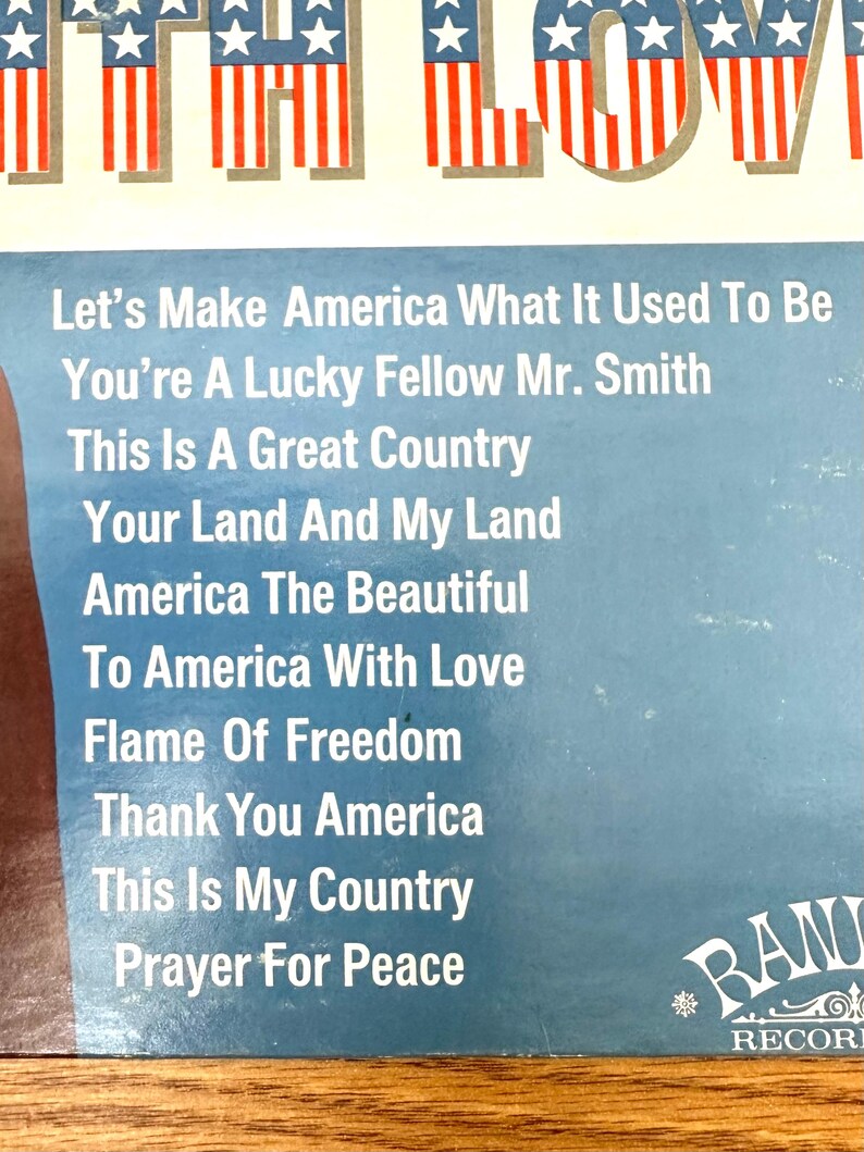 Op de afbeelding: Een vinylplatenhoes met een blauwe achtergrond en een lijst met songtitels: Let's Make America What It Used To Be, You're A Lucky Fellow Mr. Smith, This Is A Great Country, Your Land And My Land, America The Beautiful, To America With Love, Flame Of Freedom, Thank You America, This Is My Country, Prayer For Peace. De bovenrand toont een rood, wit en blauw Amerikaans vlagpatroon.