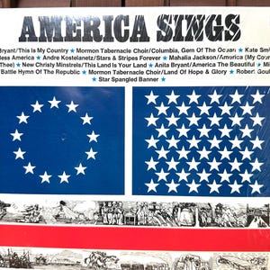 Op de afbeelding: Een vinylplatenhoes met de titel America Sings met een tracklist van patriottische liedjes. Het ontwerp bevat twee blauwe rechthoeken met witte sterren, een horizontale rode streep en zwart-wit historische illustraties met gele franje.