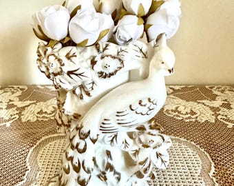 May include: A white ceramic vase featuring a sculpted peacock with gold-painted accents and floral relief details. The vase is filled with a cluster of white artificial roses with green stems, creating a decorative arrangement with a glossy finish.