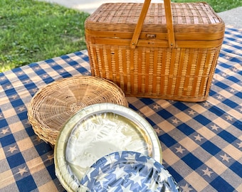 May include: A rectangular woven wicker picnic basket with a hinged lid and a central handle, accompanied by a round woven wicker tray and two stacked disposable plates, one with a blue and white star pattern, wrapped in clear plastic film.