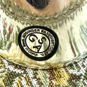 Pode incluir: Close-up de um objeto prateado com um autocolante a preto e branco que diz "Godinger Silver Art Co. Ltd. Made in China". O objeto est&aacute; sobre uma superf&iacute;cie estampada com tons de verde, amarelo e rosa.