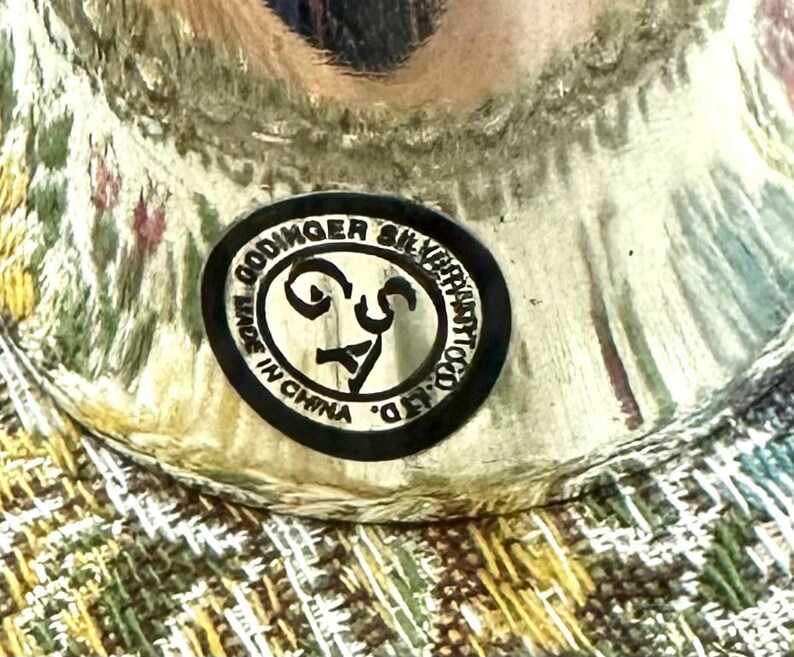 Pode incluir: Close-up de um objeto prateado com um autocolante a preto e branco que diz "Godinger Silver Art Co. Ltd. Made in China". O objeto est&aacute; sobre uma superf&iacute;cie estampada com tons de verde, amarelo e rosa.