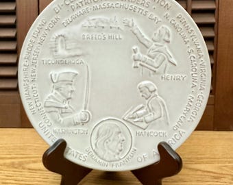 May include: A round white ceramic commemorative plate featuring embossed relief designs of historical figures including Washington, Henry, Hancock, and Benjamin Franklin, with text reading Bi-Centennial Celebration and United States of America.