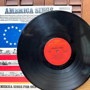 Op de afbeelding: Een zwarte vinylplaat met een rood label in het midden getiteld America Sings van Columbia Special Products. De plaat ligt voor de albumhoes, die een blauw veld met witte sterren en rode en witte strepen bevat.