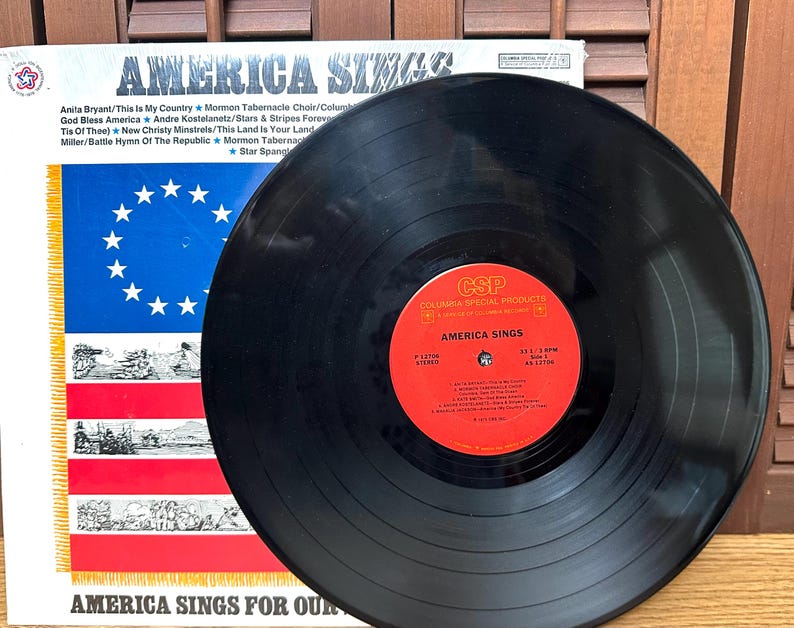 Op de afbeelding: Een zwarte vinylplaat met een rood label in het midden getiteld America Sings van Columbia Special Products. De plaat ligt voor de albumhoes, die een blauw veld met witte sterren en rode en witte strepen bevat.