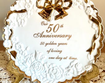May include: A white ceramic decorative plate with a scalloped edge and gold trim. The plate features embossed white roses and a central gold bow. Text reads: Our 50th Anniversary, 50 golden years of loving one day at time. It rests on a gold stand.