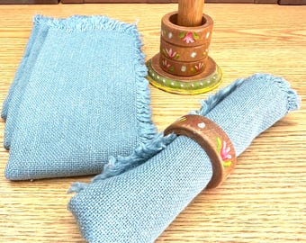 May include: A set of light blue fabric napkins with frayed edges, accompanied by a wooden napkin holder stand and matching wooden napkin rings. The rings and the base of the stand feature hand-painted pink floral motifs and white dots.