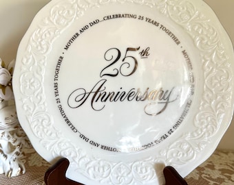 May include: A white ceramic decorative plate with a scalloped, embossed border. The centre features the text 25th Anniversary in silver lettering, surrounded by the repeating phrase MOTHER AND DAD...CELEBRATING 25 YEARS TOGETHER in black.