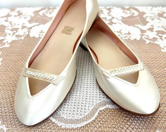May include: A pair of cream-coloured satin ballet flats featuring a pointed toe and a decorative strap across the vamp adorned with a row of small clear crystals. The interior insole is stamped with the text Comfy Slippers in a rectangular box.