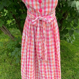 K&ouml;nnte beinhalten: Ein rosa-wei&szlig;es kariertes, &auml;rmelloses Kleid mit Schleife in der Taille. Das Kleid hat f&uuml;nf rosa Kn&ouml;pfe auf der R&uuml;ckseite.