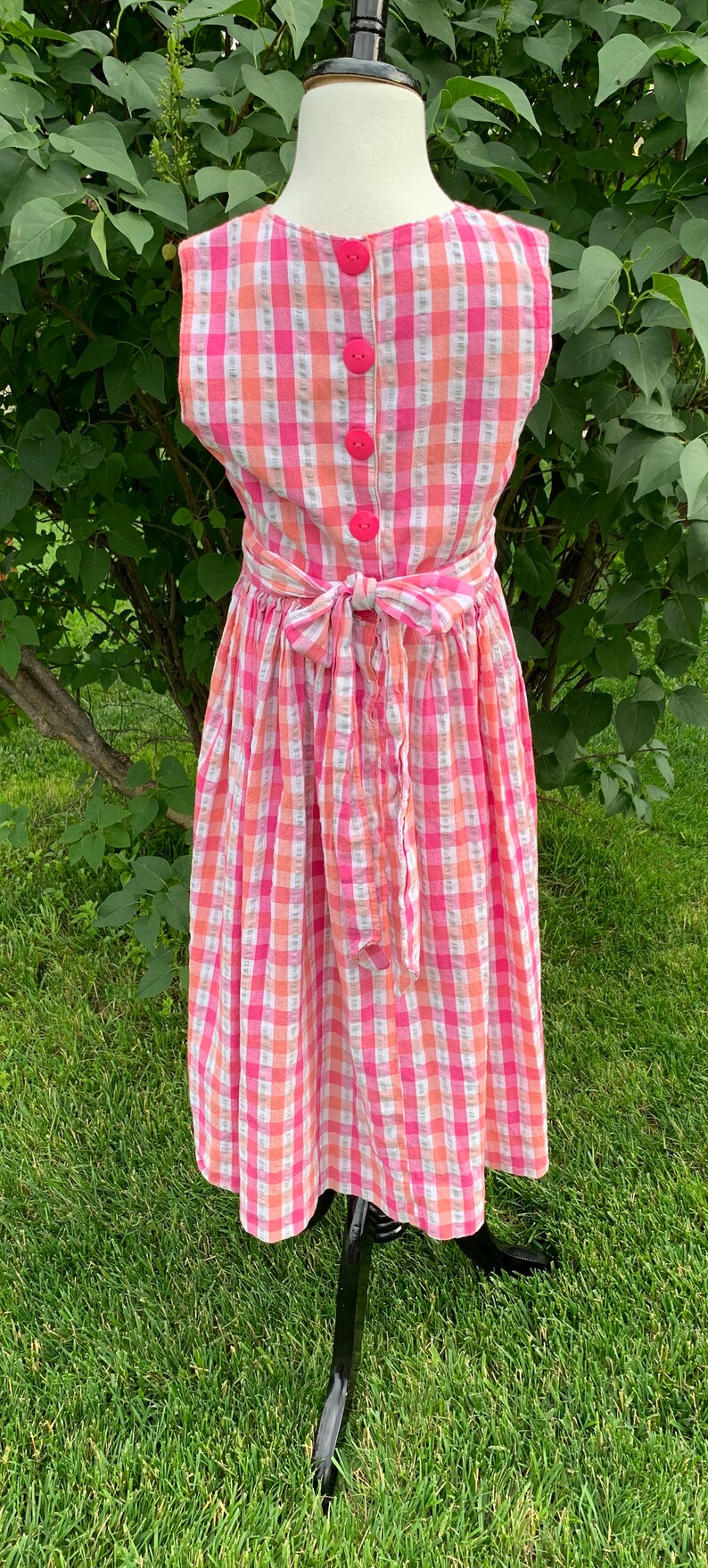 K&ouml;nnte beinhalten: Ein rosa-wei&szlig;es kariertes, &auml;rmelloses Kleid mit Schleife in der Taille. Das Kleid hat f&uuml;nf rosa Kn&ouml;pfe auf der R&uuml;ckseite.