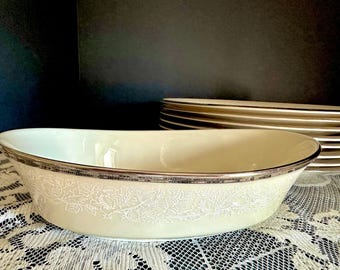 May include: An oval-shaped cream-coloured serving bowl with a metallic silver rim and a delicate white floral pattern etched around the exterior. The bowl is positioned in front of a stack of matching cream-coloured plates with silver trim.