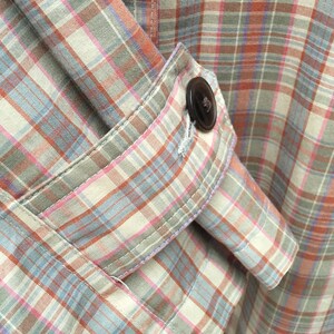 May include: A close-up of a plaid shirt with a button on the sleeve. The shirt is made of a soft, lightweight fabric and has a relaxed fit.