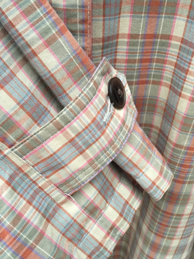 May include: A close-up of a plaid shirt with a button on the sleeve. The shirt is made of a soft, lightweight fabric and has a relaxed fit.