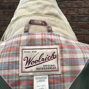 May include: A green jacket with a plaid lining and a Woolrich label. The label says "Since 1830 Woolrich Original Outdoorwear" and "Womens Large 100% Cotton Made in Thailand".