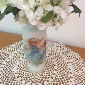 May include: White ceramic vase with a floral design featuring two girls in a field of flowers. The vase is filled with white flowers.