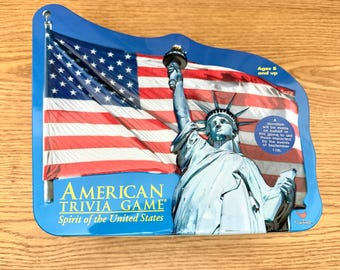 May include: A shaped metal tin for an American Trivia Game featuring the Statue of Liberty and a large American flag. The text reads American Trivia Game Spirit of the United States, Ages 8 and up, and a note about a donation for September 11th.