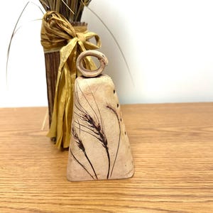Peut inclure: Une cloche en céramique beige avec un motif d'épi de blé. La cloche a une petite boucle en haut et est décorée d'épis de blé bruns. Un bouquet de blé séché et un vase avec un ruban jaune sont en arrière-plan.