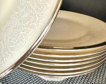 May include: A stack of round ceramic plates with a cream-coloured surface and a metallic silver rim. The top plate features a white floral pattern along the edge, while the other plates in the stack show a consistent metallic silver trim.