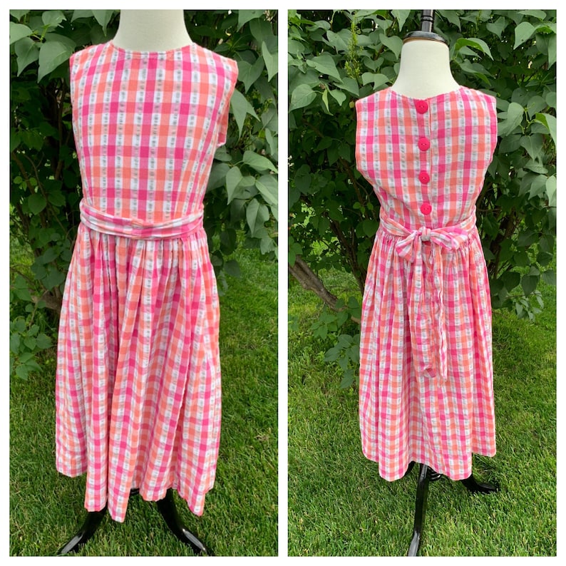 K&ouml;nnte beinhalten: Ein rosa-wei&szlig;es kariertes, &auml;rmelloses Kleid mit Schleife in der Taille und Kn&ouml;pfen im R&uuml;cken.