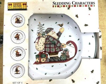 Könnte beinhalten: Set mit vier weißen Steingut-Salat- oder Desserttellern mit einer Winterszene, in der der Weihnachtsmann Schlitten fährt. Die Teller sind mit einem rot-grünen Karomuster, einem Weihnachtsbaum und einem Vogel verziert. Die Schachtel trägt die Aufschrift "Debbie Mumm's Word" und "Sledding Characters".
