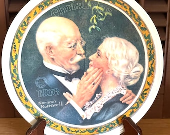 May include: A round ceramic decorative plate featuring a Norman Rockwell illustration of an elderly couple under mistletoe. The plate displays the text "Christmas", "1976", and "Norman Rockwell" with a yellow and green holly leaf border design.