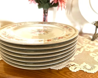 May include: A stack of eight white ceramic dinner plates with a floral pattern in shades of pink, yellow, and green. The plates have a gold trim around the edge.