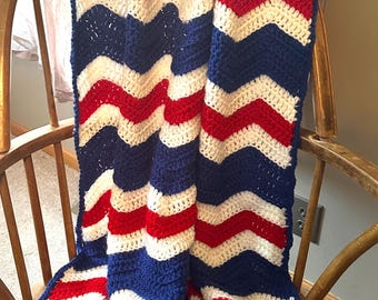 May include: A crocheted blanket featuring a repeating chevron pattern in solid stripes of royal blue, white, and bright red. The textile has a textured, yarn-based construction with a consistent wave design that runs vertically throughout the entire piece.