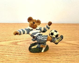 May include: A brown teddy bear figurine wearing a blue and white striped football kit, kicking a black and white football. The bear is positioned on a textured green base, captured in a dynamic motion with one leg extended forward.