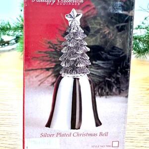 Pode incluir: Um sino de Natal prateado com um design de &aacute;rvore de Natal encimado por uma estrela. O sino tem uma base branca com listras pretas e uma borda vermelha. A caixa diz "Holiday Collection by Godinger".