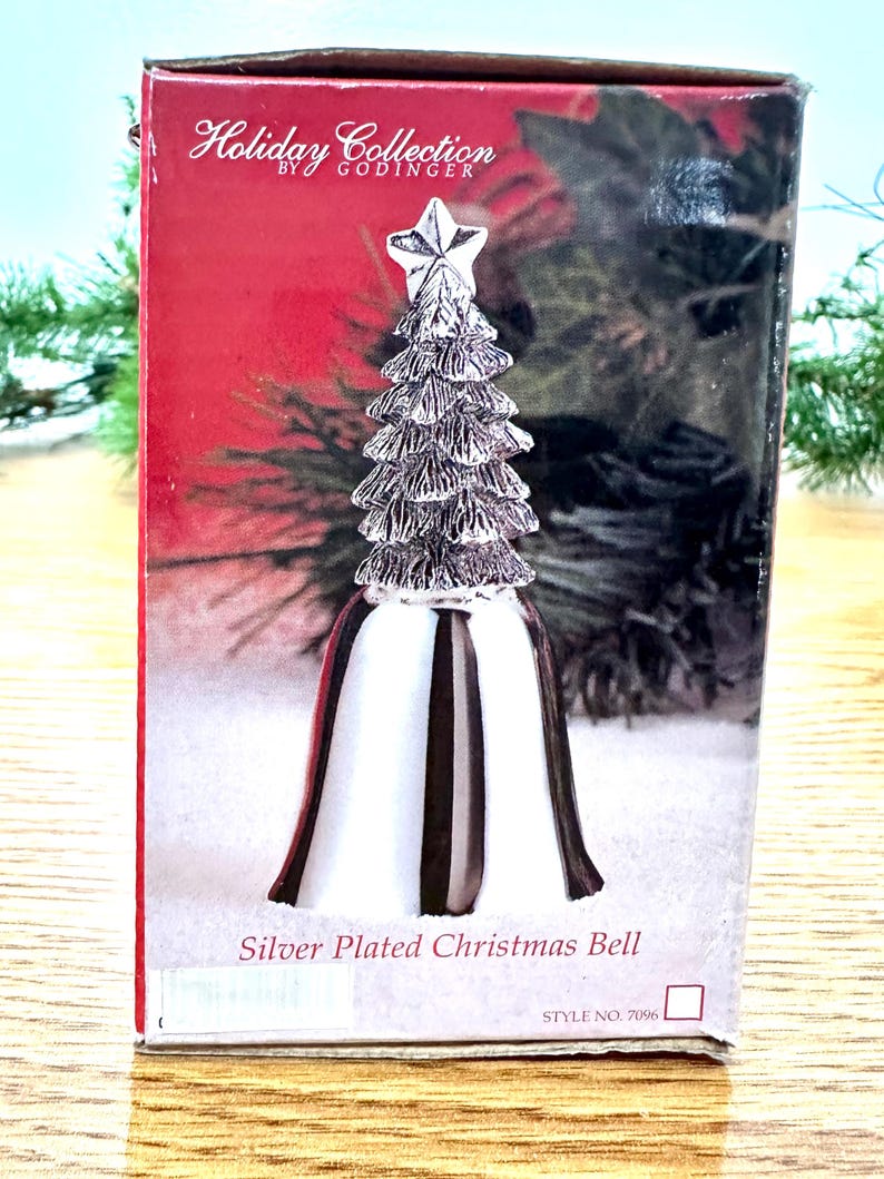Pode incluir: Um sino de Natal prateado com um design de &aacute;rvore de Natal encimado por uma estrela. O sino tem uma base branca com listras pretas e uma borda vermelha. A caixa diz "Holiday Collection by Godinger".