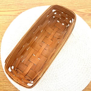 May include: A woven wooden basket with a rectangular shape and a natural brown color. The basket is empty and has a woven pattern.