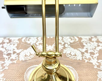 May include: A polished brass piano lamp featuring a wide, horizontal, adjustable shade with a white interior and a single tubular light bulb. The lamp has a sturdy, round base and a vertical arm with a central hinge for height adjustment.