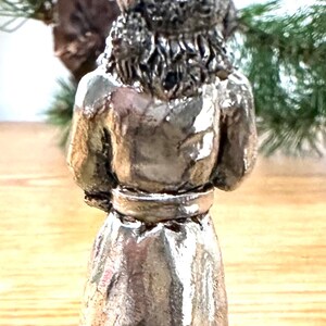 Pode incluir: Estatueta prateada de uma pessoa, vista por tr&aacute;s. A figura usa uma t&uacute;nica longa com cinto e chap&eacute;u. A base da estatueta tem um design vazado decorativo. O fundo inclui vegeta&ccedil;&atilde;o.
