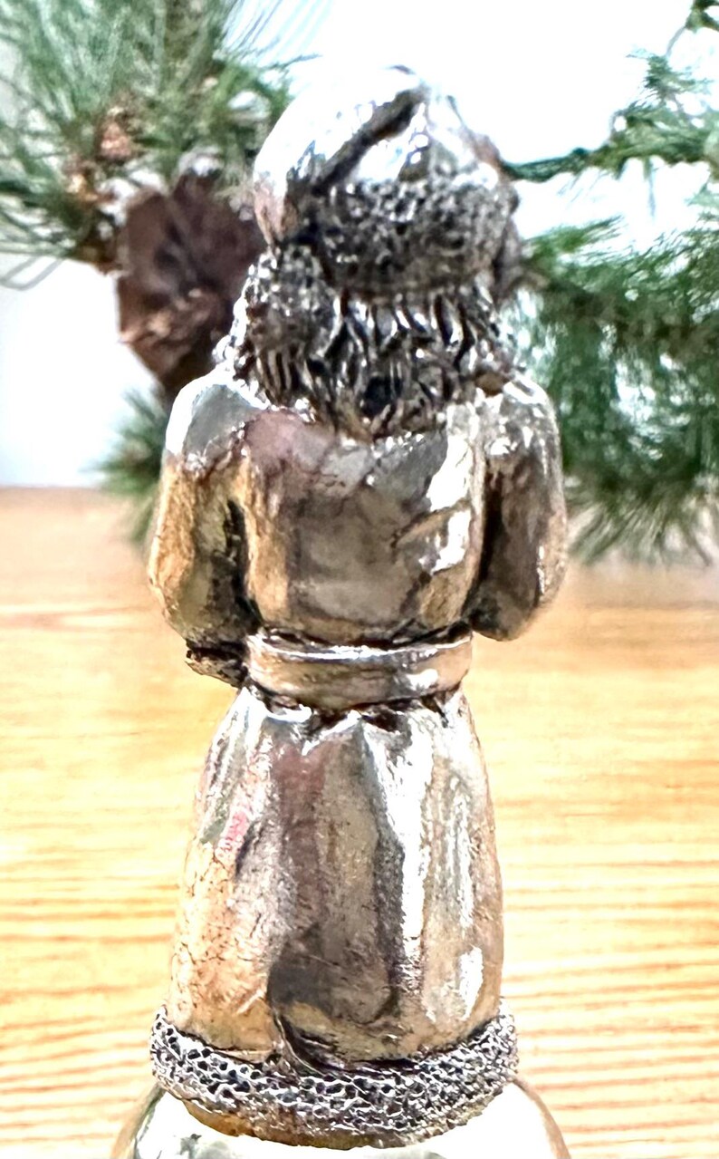 Pode incluir: Estatueta prateada de uma pessoa, vista por tr&aacute;s. A figura usa uma t&uacute;nica longa com cinto e chap&eacute;u. A base da estatueta tem um design vazado decorativo. O fundo inclui vegeta&ccedil;&atilde;o.