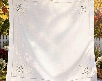 May include: A square white linen tablecloth featuring delicate cutwork embroidery in each corner. The design includes intricate floral patterns and geometric shapes created with white thread, framed by a simple stitched border along the edges.