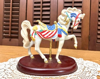 May include: A white carousel horse figurine featuring a saddle decorated with blue and white stars and red and white stripes. The horse has a flowing mane, a gold-colored pole, and stands on a dark brown oval wooden base with gold-painted hooves.