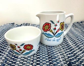 May include: A white ceramic creamer and a matching small bowl, both decorated with colorful floral patterns in red, yellow, and blue. The creamer features the text "Var så god" in blue script, a pouring spout, and a handle on the side.