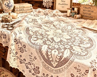 May include: A white lace tablecloth featuring an intricate floral and leaf pattern with a scalloped border. The fabric has a delicate openwork design and is draped over a surface. The lace displays a central medallion motif surrounded by floral vines.