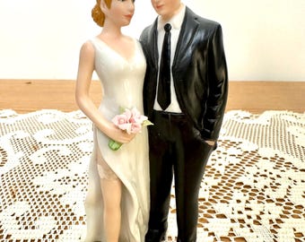 May include: A ceramic wedding cake topper figurine featuring a person in a white sleeveless dress with a high leg slit and a garter, holding a small bouquet of pink flowers, standing next to a person in a black suit with a white shirt and black tie.
