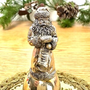 Pode incluir: Estatueta de sino do Papai Noel prateada. A figura do Papai Noel &eacute; detalhada com um chap&eacute;u, barba e casaco texturizados, segurando um saco. O sino tem uma superf&iacute;cie polida e reflexiva. Decora&ccedil;&atilde;o de Natal festiva.
