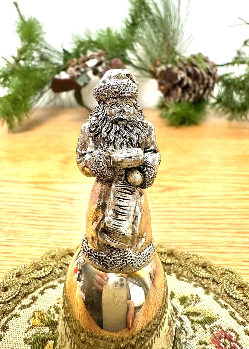 Pode incluir: Estatueta de sino do Papai Noel prateada. A figura do Papai Noel &eacute; detalhada com um chap&eacute;u, barba e casaco texturizados, segurando um saco. O sino tem uma superf&iacute;cie polida e reflexiva. Decora&ccedil;&atilde;o de Natal festiva.