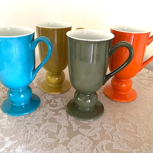 May include: Four ceramic mugs with different coloured glazes. The mugs have a footed base and a handle. The colours are teal, orange, green, and yellow.