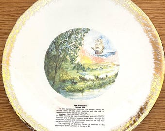 May include: A round white ceramic plate with a gold-trimmed edge featuring a central circular illustration of a sailing ship, a horse-drawn wagon, and a landscape. Below the image, black text reads: New Scandinavia Settled 1868, followed by a paragraph describing the pioneers' journey to Kansas, noting the painting is by Esther Johnson.