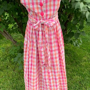 K&ouml;nnte beinhalten: Ein rosa-wei&szlig;es kariertes, &auml;rmelloses Kleid mit Schleife in der Taille. Das Kleid hat vier rosa Kn&ouml;pfe auf dem R&uuml;cken.