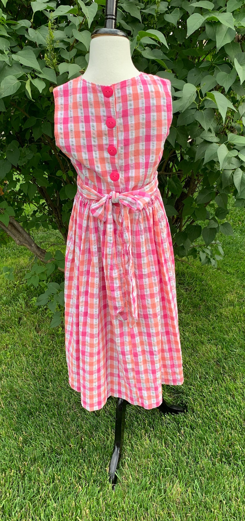 K&ouml;nnte beinhalten: Ein rosa-wei&szlig;es kariertes, &auml;rmelloses Kleid mit Schleife in der Taille. Das Kleid hat vier rosa Kn&ouml;pfe auf dem R&uuml;cken.