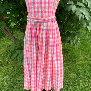 K&ouml;nnte beinhalten: Ein &auml;rmelloses Kleid in rosa und wei&szlig;em Gingham mit einem passenden G&uuml;rtel. Das Kleid befindet sich auf einer Puppe und wird vor einem gr&uuml;nen Gras Hintergrund pr&auml;sentiert.